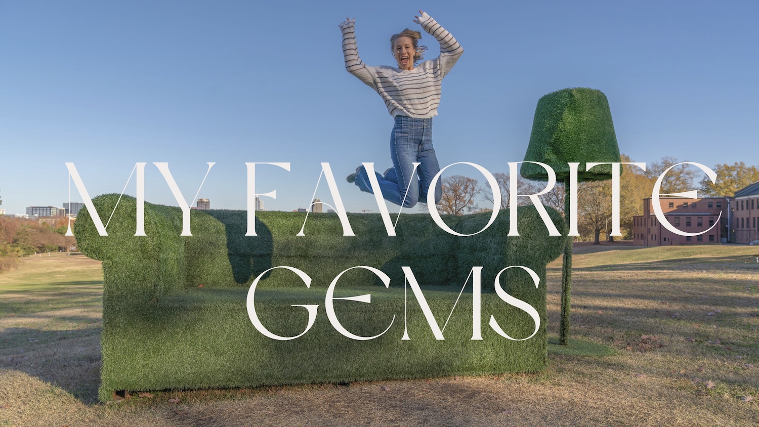 Erika jumping for joy on an oversized grass sofa sculpture at Dix Park, with the Raleigh skyline behind her