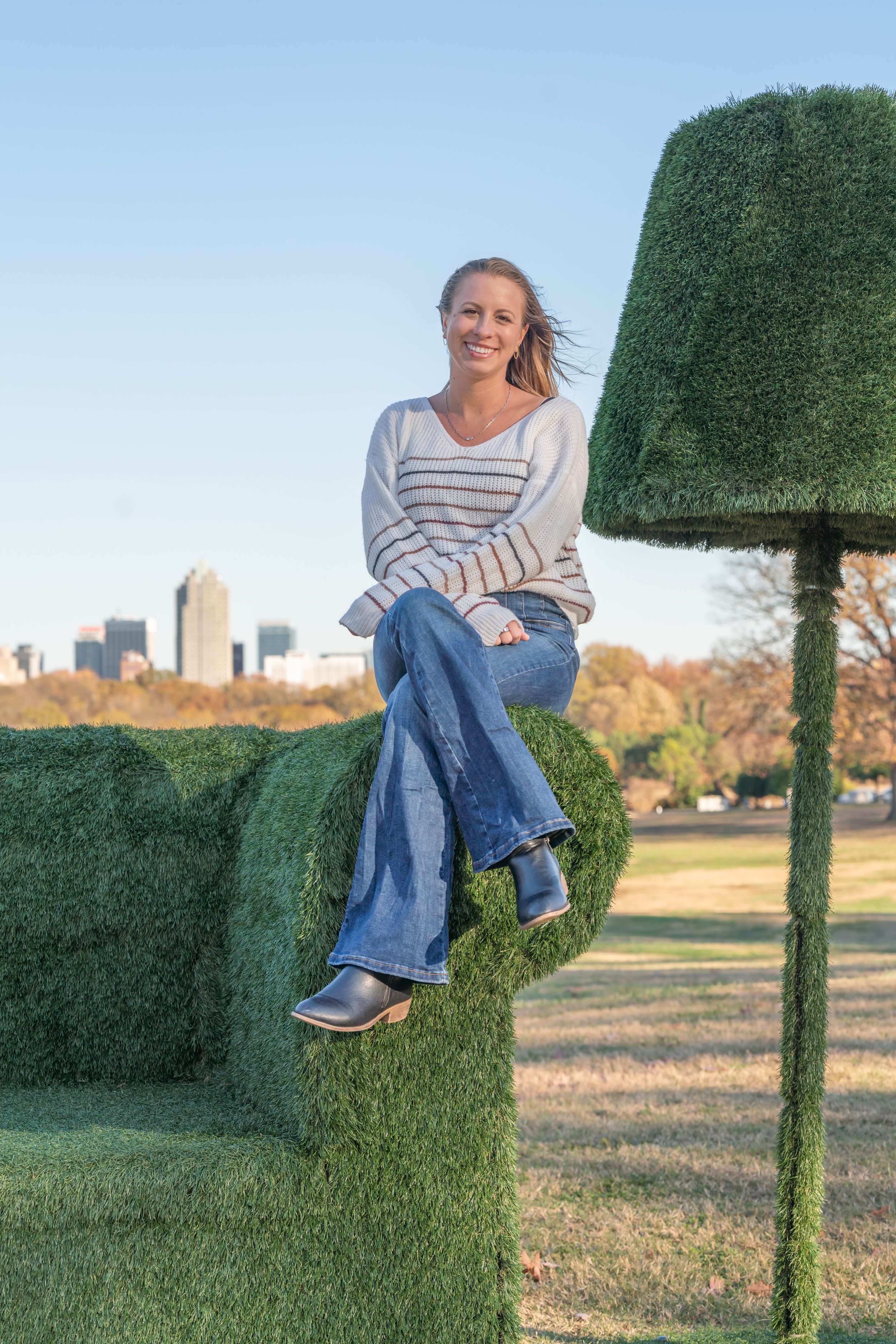 Erika sitting on the grass sofa sculpture at Dix Park with the Raleigh skyline in the background